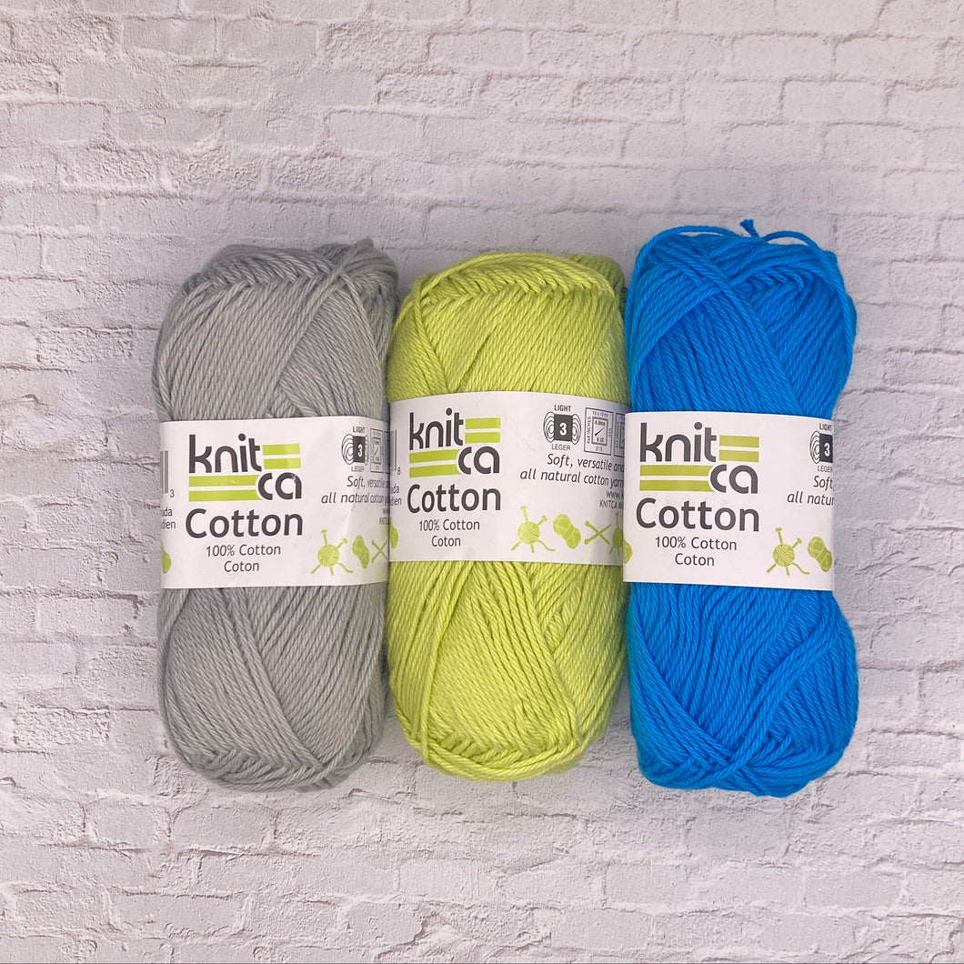 Three skeins of knit cotton yarn in gray, green, and blue on a white brick wall background.